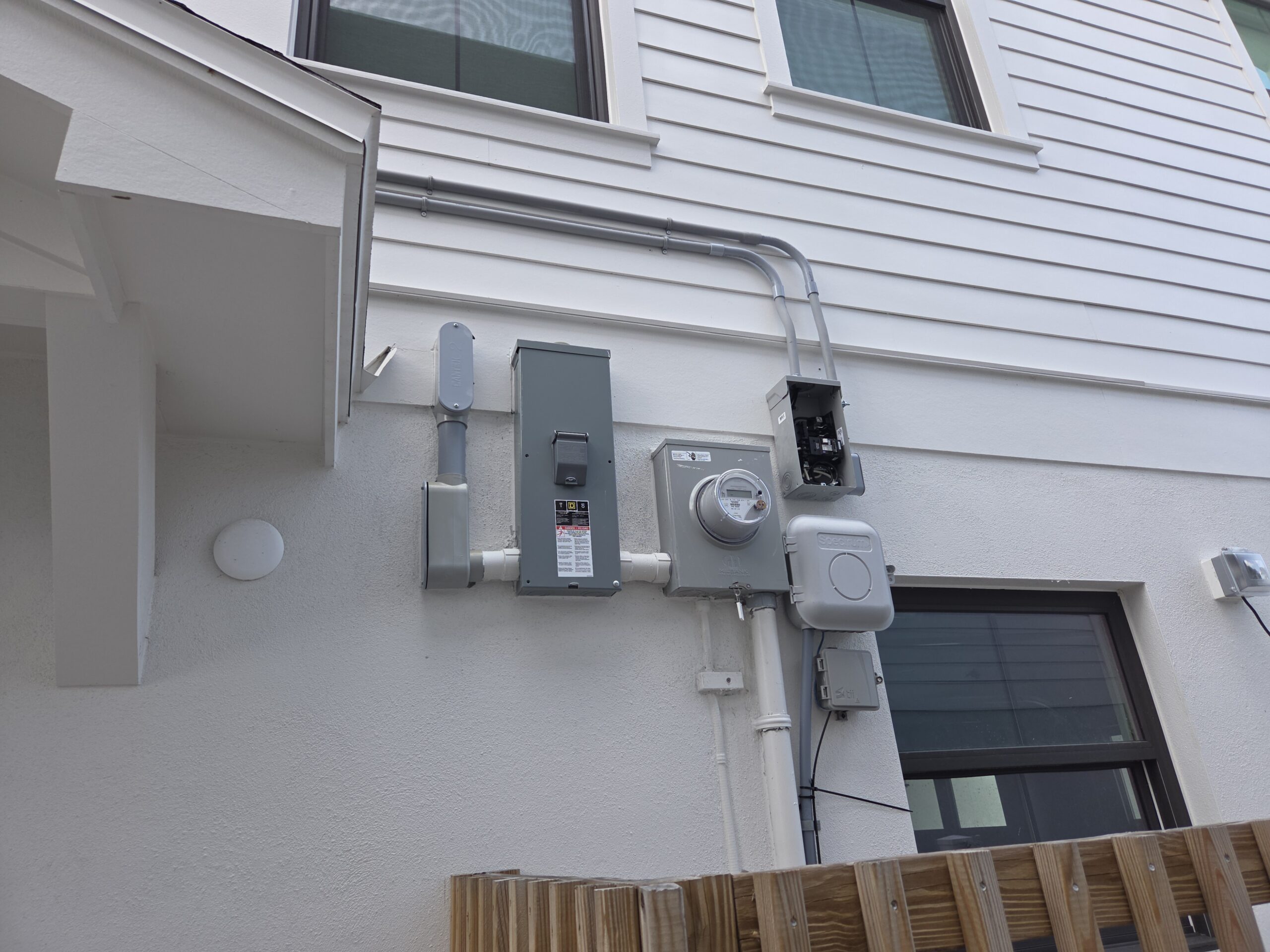 Exterior house wall in Shore Acres, St. Petersburg with electrical meter box, conduit pipes, and utility panels beneath two windows. This includes an electrical subpanel relocation.
