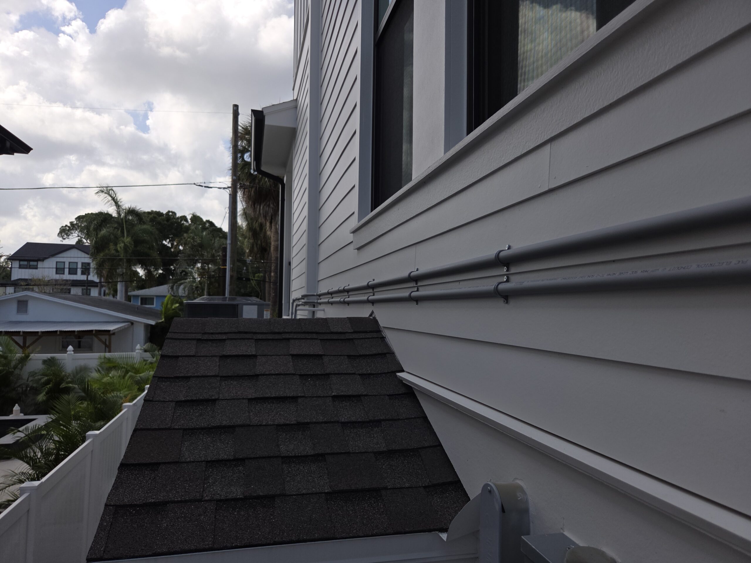 Exterior view of a light-gray siding house with a window and metal gutters, palm trees and power lines visible in the background. Electrical conduit running outside the house.